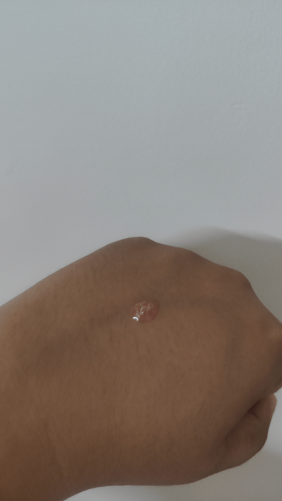A close-up image of a hand with a drop of clear serum sitting on the skin against a plain white background.